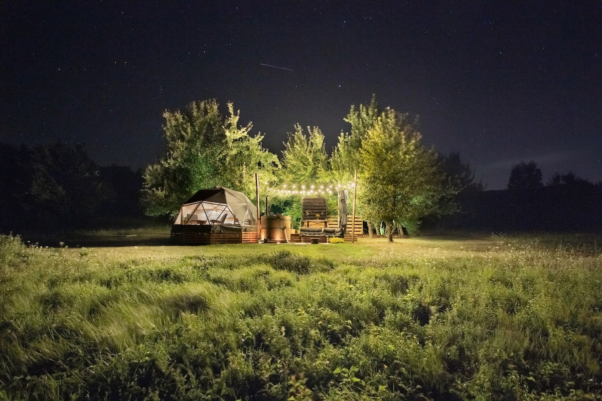 A glamping site is set against a night sky, with warm string lights illuminating a wooden terrace surrounded by trees. A dome structure is visible nearby, blending with the natural landscape of grassy fields.