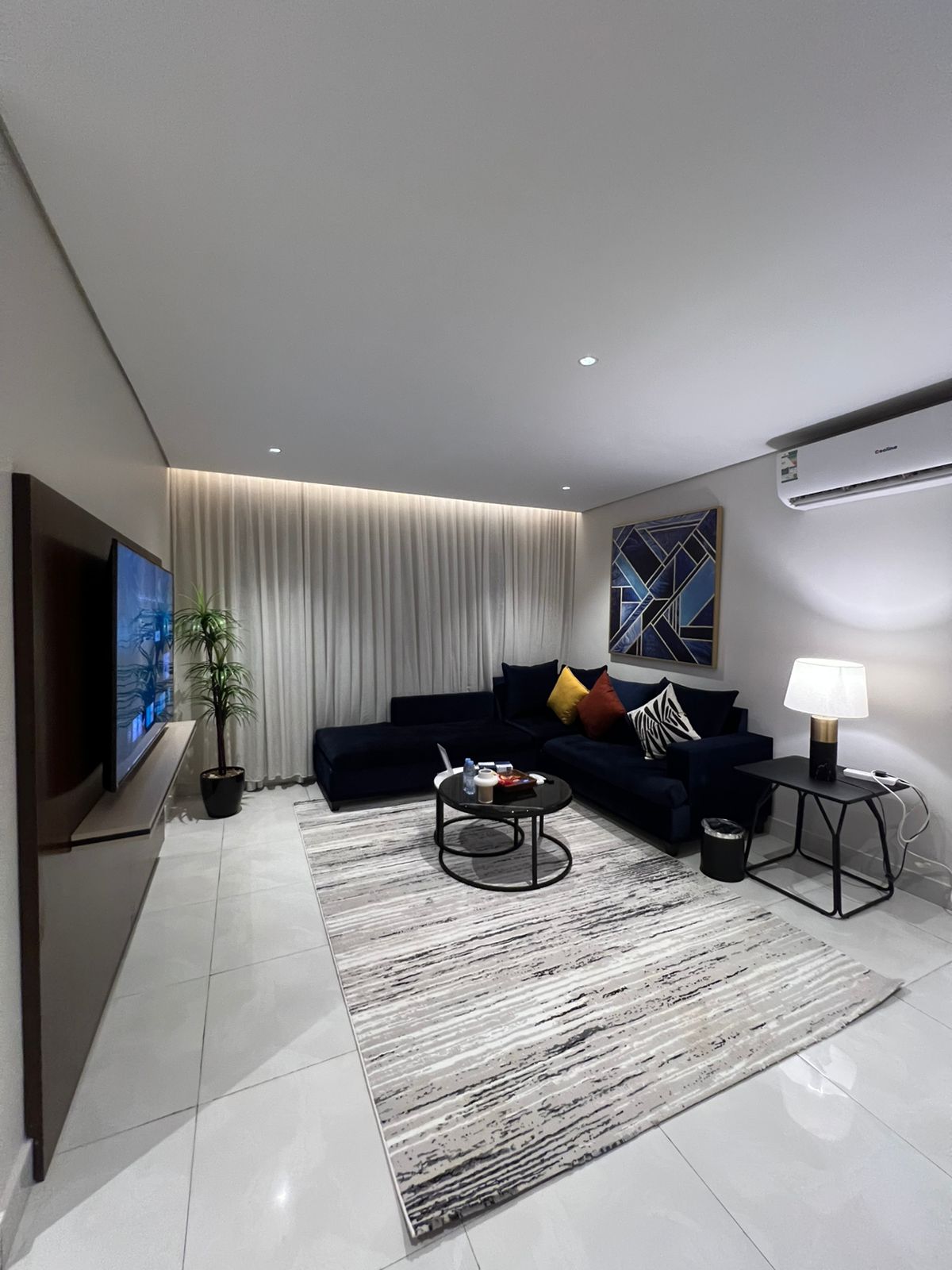 A modern living area is presented, featuring a dark blue sectional sofa with colorful cushions. A round coffee table sits on a light rug, while a potted plant adds greenery. Soft lighting and an air conditioning unit are visible, enhancing comfort.