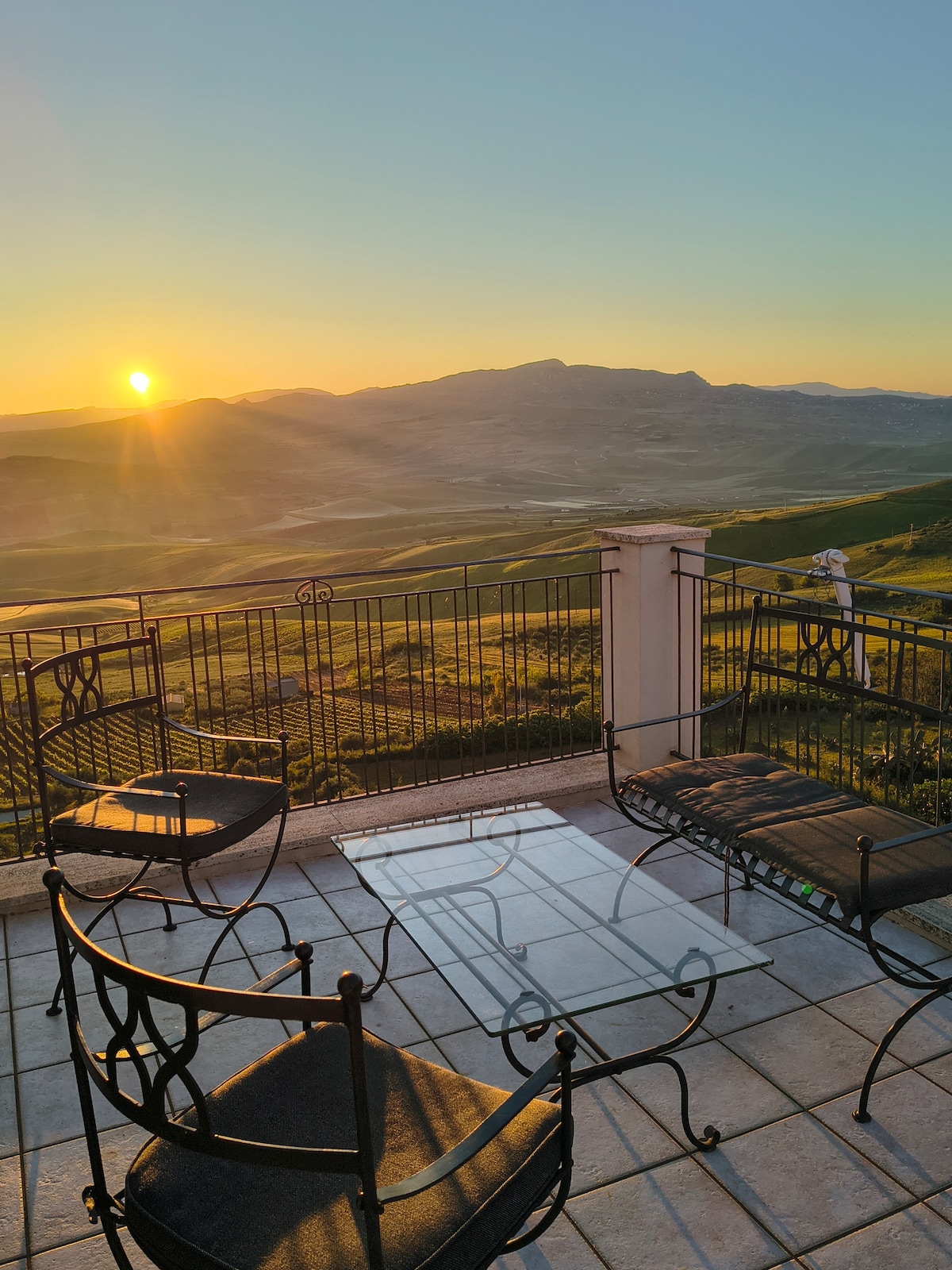 A terrace is showcased with a glass table and four metal chairs, capturing an expansive view of rolling hills bathed in sunlight. The panoramic landscape is enhanced by the warm glow of the setting sun, while a gentle breeze can be imagined flowing through the peaceful space.