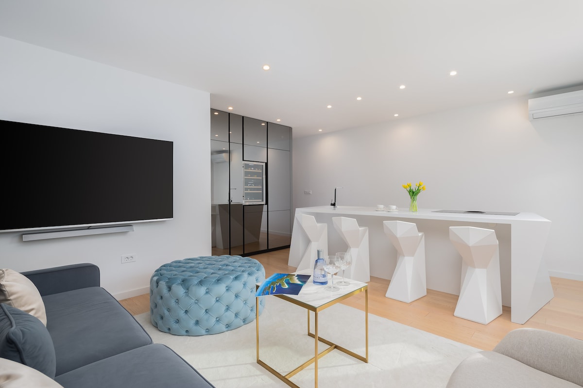 A modern living space features sleek white cabinetry and a central island with bar stools. A plush blue ottoman complements a gray sofa, while light fixtures above provide illumination. A large television is mounted on the wall opposite the seating area, enhancing the contemporary design.
