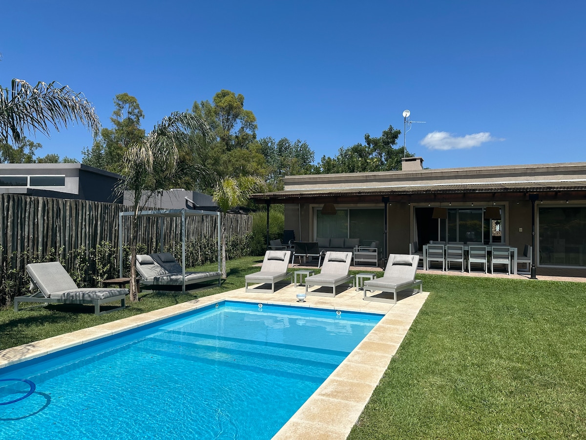 A serene outdoor space is presented, featuring a clear blue swimming pool surrounded by sun loungers and lush green grass. A shaded area is available, with a large dining table positioned nearby. Palm trees add a touch of nature to the tranquil setting.