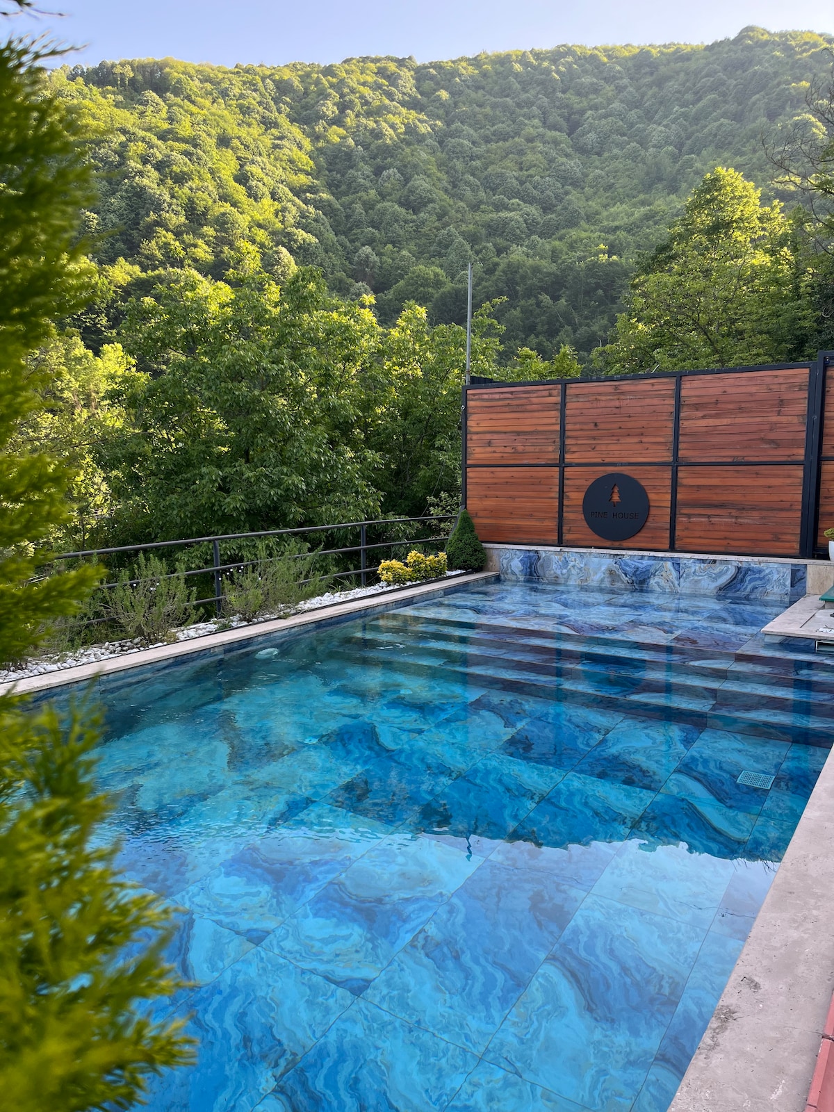 A serene outdoor pool is framed by lush greenery and wooden fencing, reflecting the vibrant blue tiles under sunlight. The mountain backdrop provides a natural, tranquil setting, enhancing the space's peaceful ambiance.