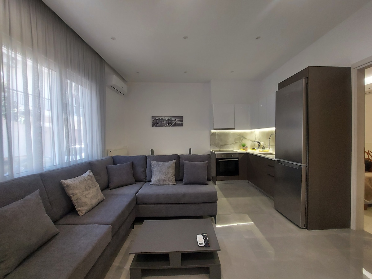 A modern living area is visible, featuring a spacious gray sectional sofa adorned with several cushions. A low coffee table sits in front of the sofa. To the right, a sleek kitchen with light-colored cabinets and modern appliances, including a stainless steel refrigerator, is showcased. Natural light filters through large windows, illuminating the space.