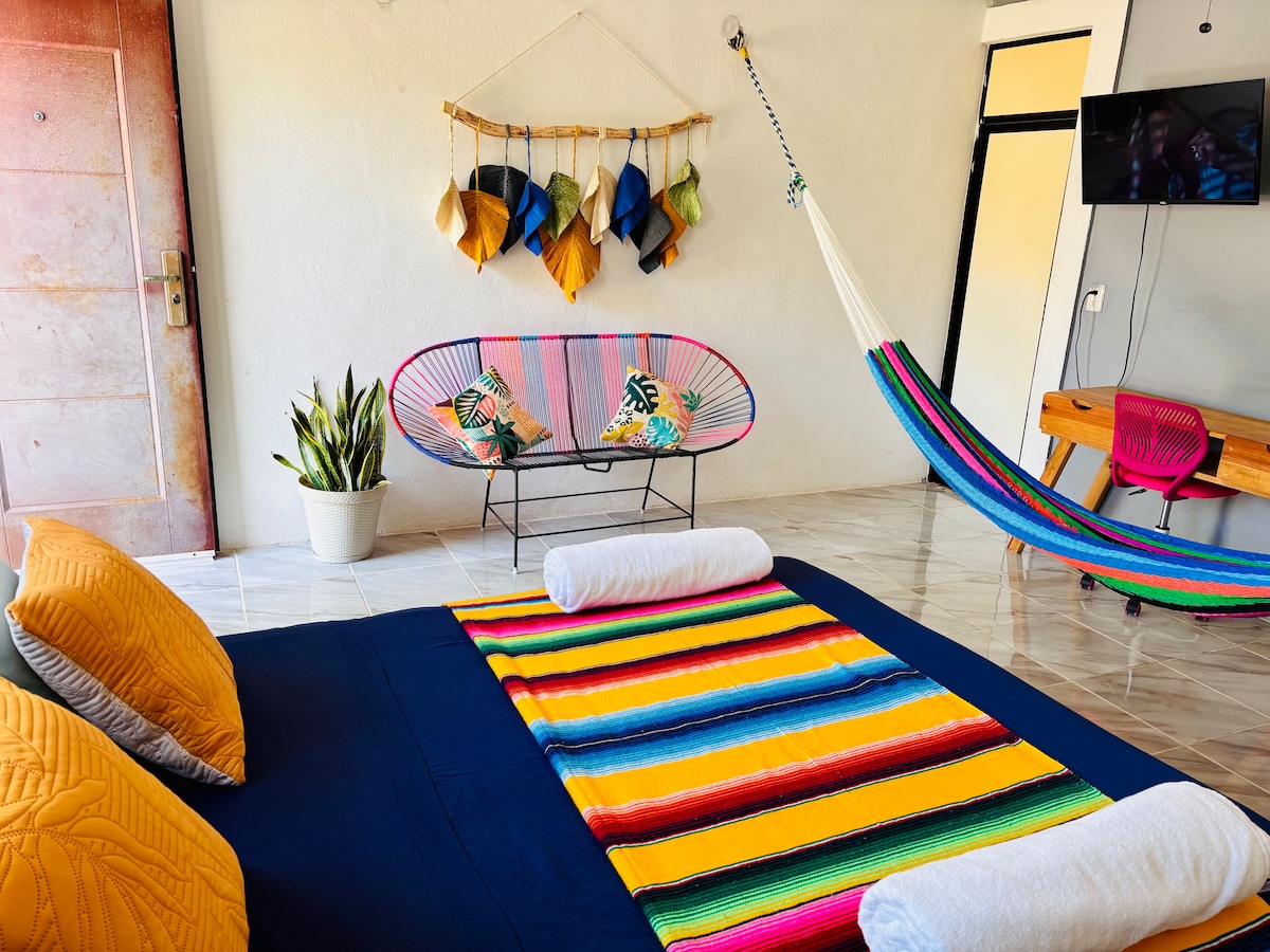A bright and spacious studio features a large bed adorned with colorful blankets. Decorative elements include wall art with hanging leaves and a hammock alongside a sleek television. Potted plants add a touch of greenery, contributing to the inviting space.