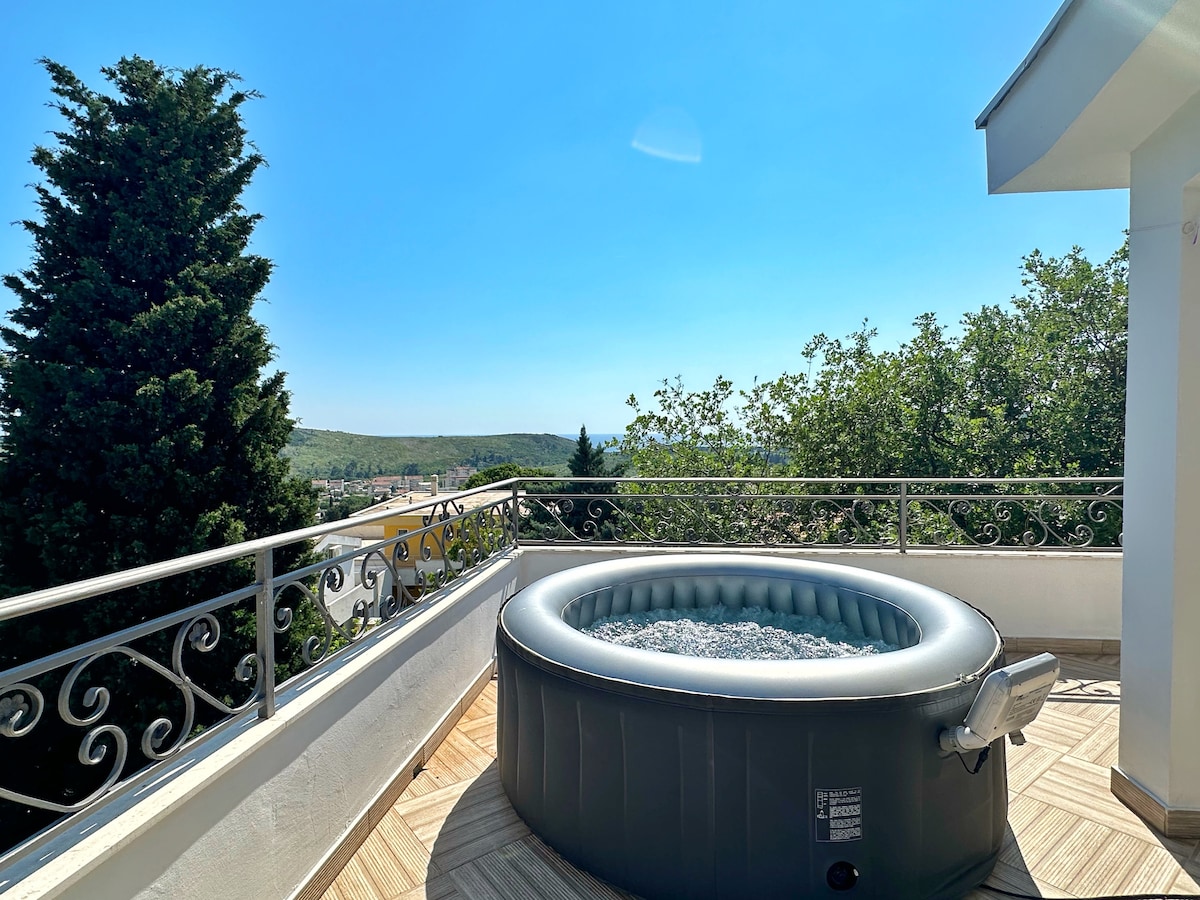 An outdoor terrace features a spacious round jacuzzi, surrounded by a wrought iron railing that offers views of greenery and hills. The clear blue sky highlights the serene environment, while the wooden flooring complements the relaxing atmosphere.
