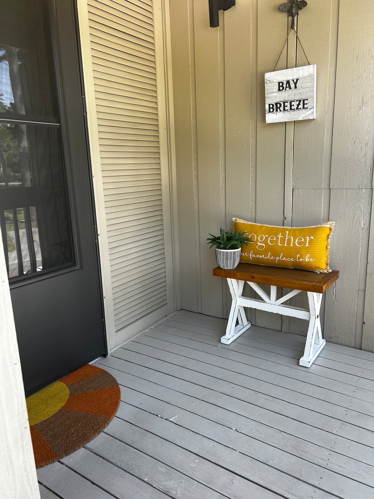 The porch area is depicted with a welcoming brown wooden bench complemented by a potted plant. A decorative yellow pillow with the word 'together' is displayed on the bench. A circular doormat adds a splash of color, while a sign reading 'BAY BREEZE' hangs on the wall.