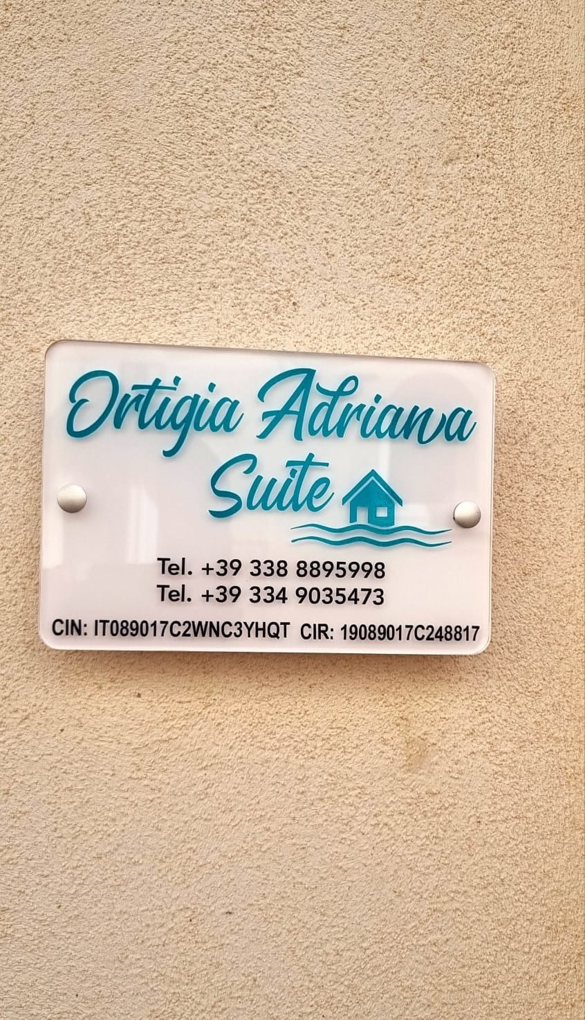 A neatly designed sign is mounted on a beige wall, displaying the name 'Ortigia Adriana Suite' in playful blue lettering. Contact numbers are listed for guests' convenience, accompanied by a simple graphic of a house and water wave.