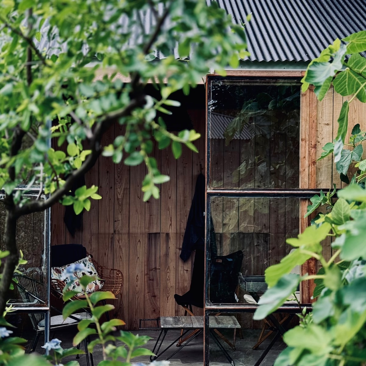 A cozy outdoor space is framed by lush greenery, featuring a wooden structure with large glass windows. Inside, minimalist seating areas are visible, providing a peaceful atmosphere. The combination of natural elements and thoughtfully designed seating enhances the inviting setting.
