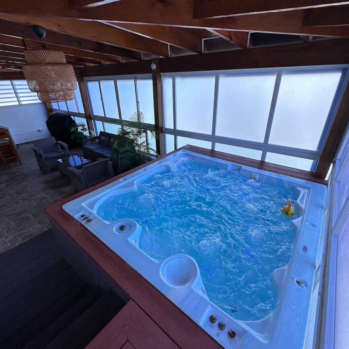 A private hot tub is featured in a well-lit area, surrounded by large windows allowing natural light. Comfortable seating is arranged nearby, creating a relaxing atmosphere. The wooden beam ceiling adds warmth to the space, enhancing the overall inviting design.