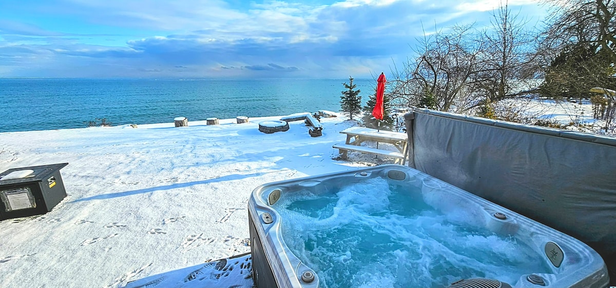 A hot tub is situated on the patio, showcasing bubbling waters against a backdrop of a serene lake and snowy landscape. Outdoor seating is visible, along with a picnic table, creating an inviting space to relax while enjoying the tranquil views.