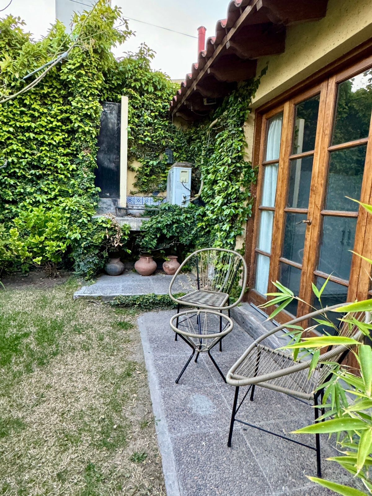 A small outdoor area is surrounded by lush greenery, featuring a stone pathway leading to a well-maintained garden. Two metal chairs are positioned on the patio, facing an ornamental wall. Natural light filters through the foliage, creating a serene atmosphere.