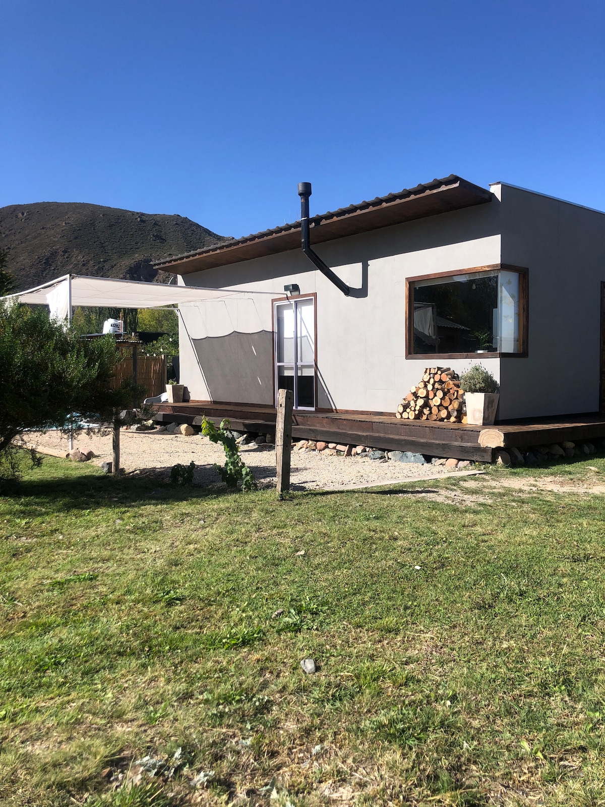 A modern cabin is positioned amidst natural surroundings, featuring a sloped roof and large windows. A wooden deck extends from the structure, accompanied by a stack of firewood and a canopy for shade. Lush greenery frames the property, set against a mountain backdrop.