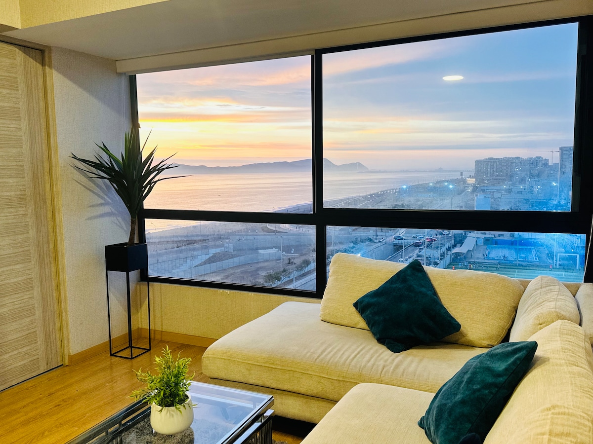 A cozy living area is situated beside a large window, showcasing a serene view of the ocean and a colorful sunset. A comfortable sectional sofa with dark green cushions is paired with a glass coffee table. A potted plant adds a touch of greenery to the space.