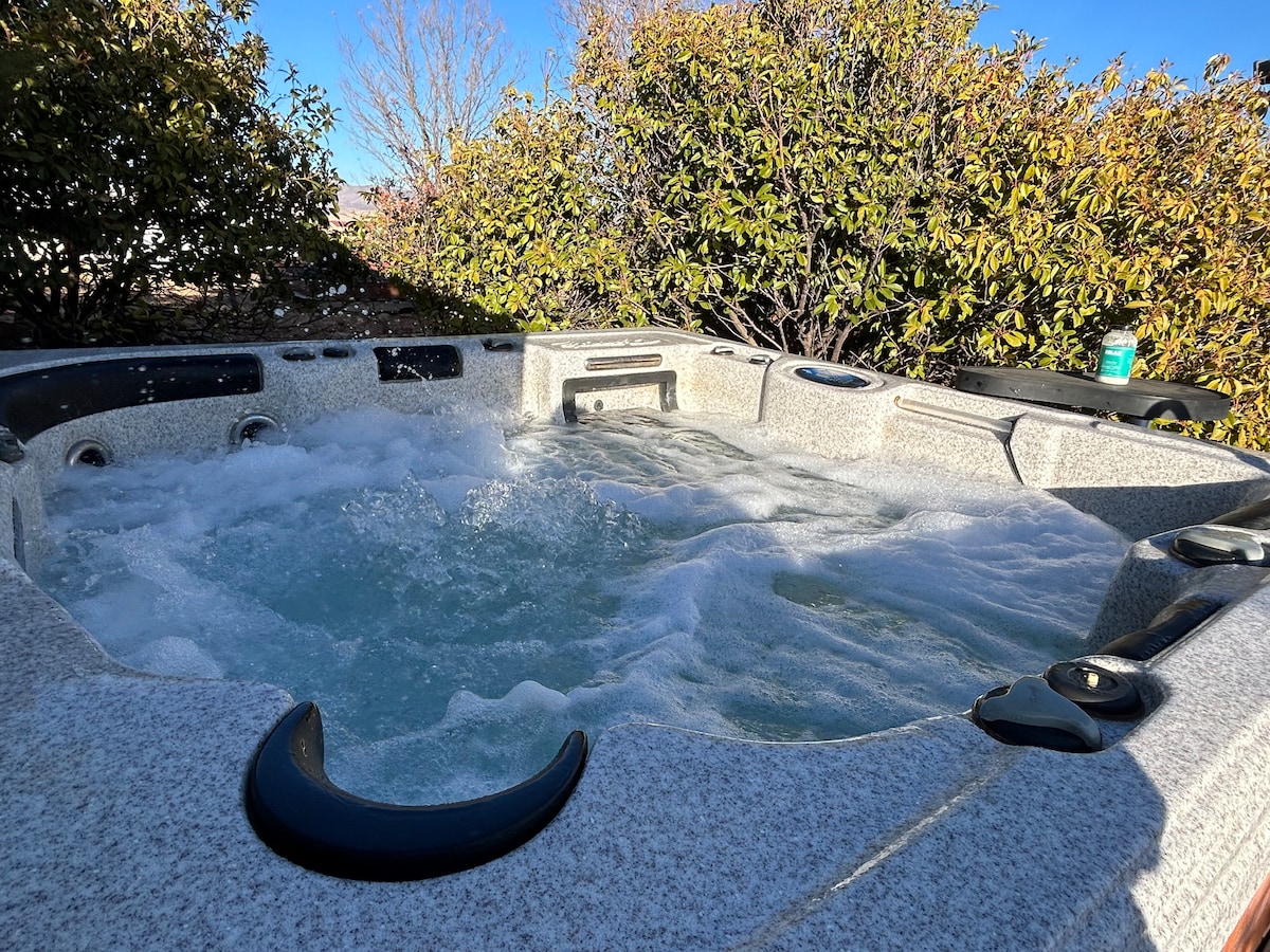 A spacious jacuzzi is surrounded by lush greenery, featuring bubbling water inviting relaxation. The surface is textured, and control panels are visible on the side, merging comfort with convenience in a tranquil outdoor setting.