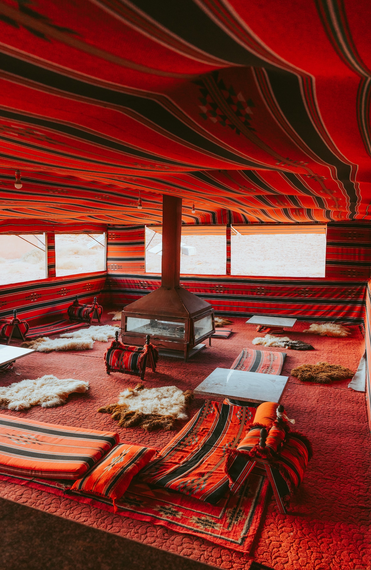 A spacious communal area showcases vibrant red and patterned textiles covering the walls and floor. A central fireplace adds warmth, surrounded by soft seating arrangements on traditional rugs. Large windows provide natural light and views of the desert landscape outside.