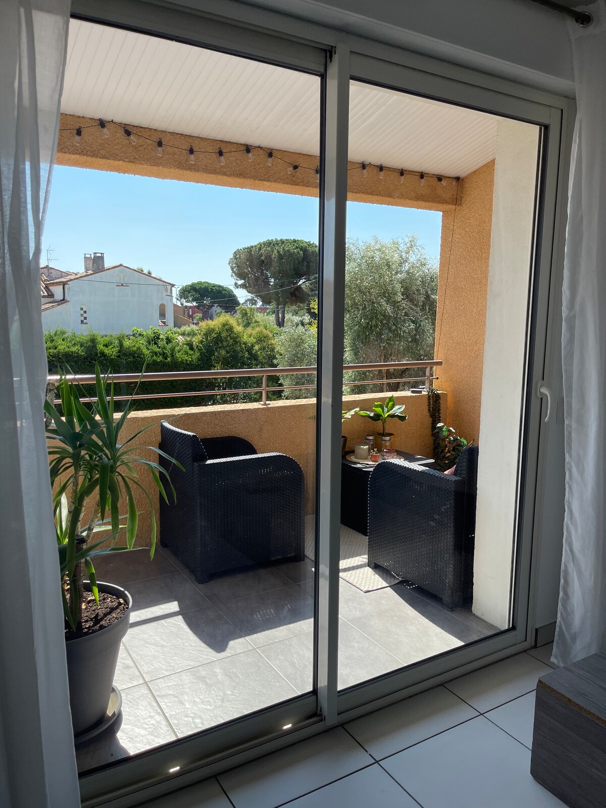 A balcony area is showcased, featuring two chairs and potted plants. Sunlight filters through sheer curtains, illuminating the space. The balcony rail offers a view of surrounding greenery, creating a serene environment for relaxation.