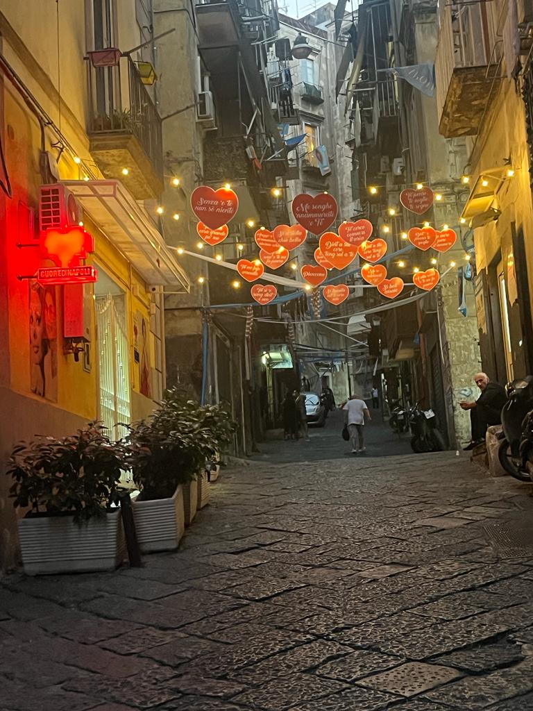 A narrow street is highlighted by strings of heart-shaped decorations illuminated by soft lights. Cobblestone pavement leads toward the far end where a pedestrian walks, flanked by buildings featuring warm-colored facades and small storefronts. Potted greenery adds a touch of nature to the scene.
