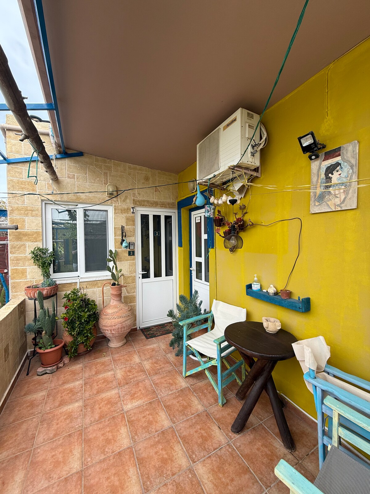 A small patio area features a round table with two chairs, surrounded by potted plants and decorative elements. A terracotta pot adds warmth, while an air conditioning unit is mounted on the wall. Light is provided by wall-mounted fixtures.