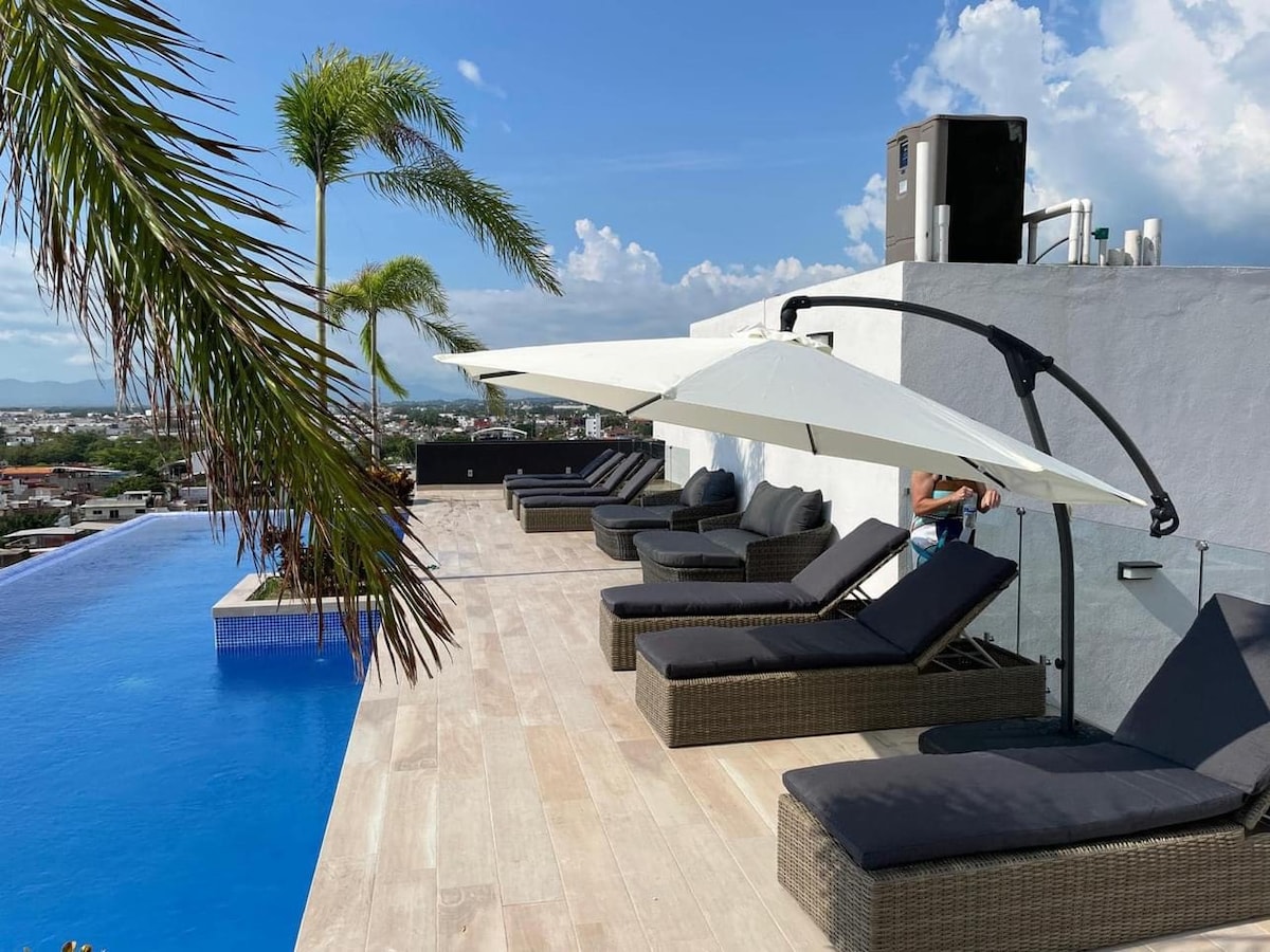 An infinity pool overlooks the town, surrounded by sun loungers beneath large umbrellas. Lush palm trees are seen in the foreground, while a clear blue sky and distant mountains provide a serene backdrop.