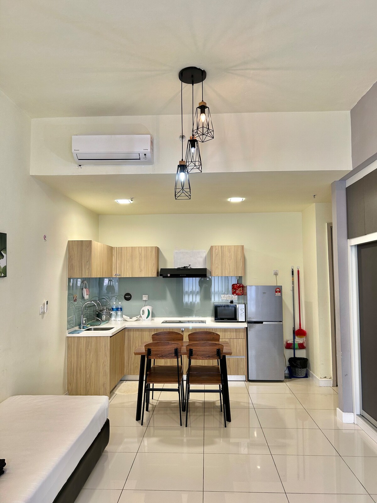 The kitchen area features modern cabinetry and appliances, including a refrigerator and microwave. A dining table for two is positioned at the forefront, complemented by a stylish light fixture overhead. The open layout is enhanced by clean tile flooring and natural light.