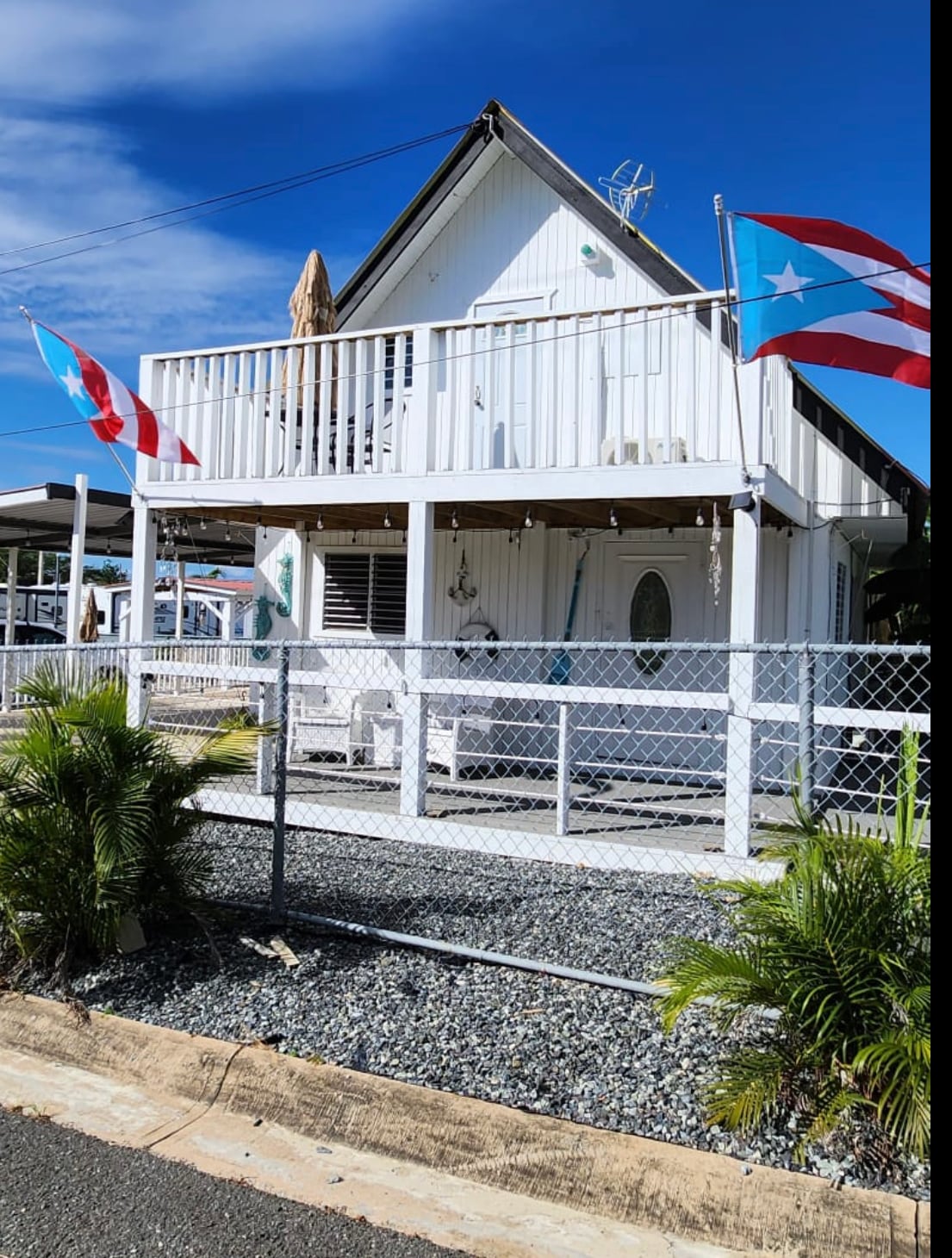 The exterior of a two-story house is depicted, featuring two balconies adorned with flags. A gated front yard with gravel and landscaped greenery surrounds the house, situated under a clear blue sky.
