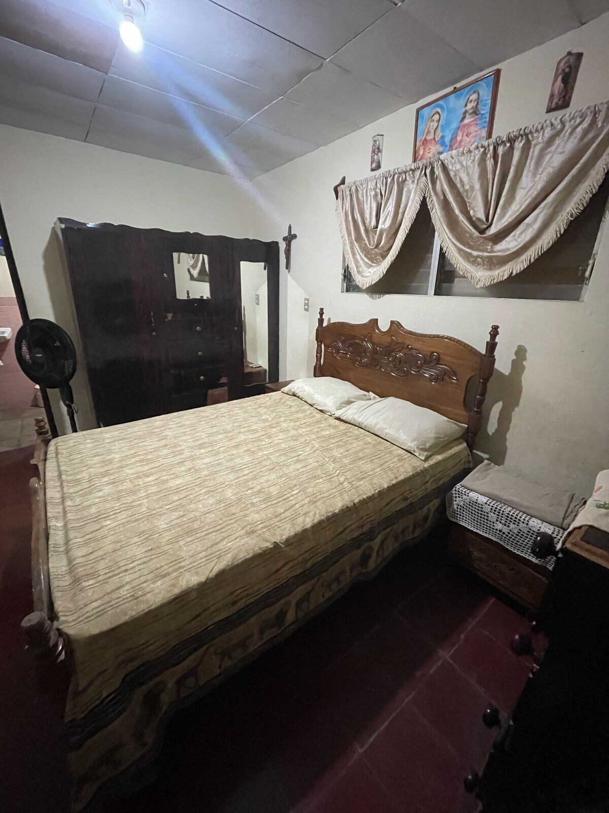 The sleeping area features a wooden bed with a patterned blanket and two pillows. A decorative mirror is displayed on a nearby cabinet. Light filters through a window covered by curtains, and religious artwork is visible on the wall.