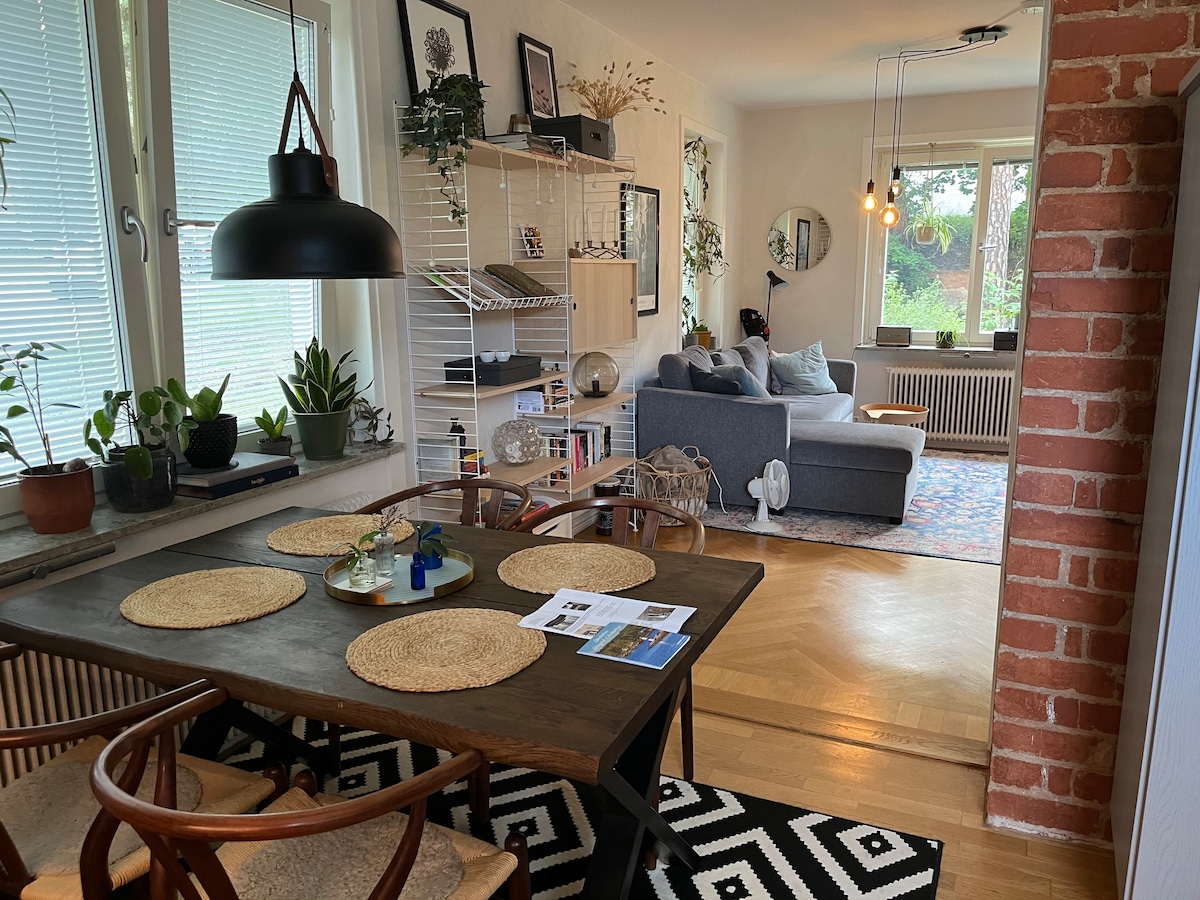 An open floor plan connects a dining area and living space. A wooden table is set with placemats and accompanied by several chairs. A cozy couch faces a window with garden views, while plants and decorative items enhance the atmosphere.