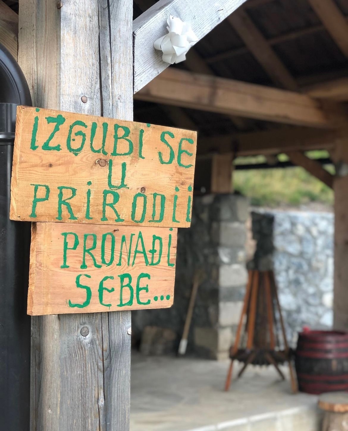 A wooden sign, featuring vibrant green lettering, promotes a message encouraging guests to connect with nature. The sign is situated under a rustic wooden structure, complemented by natural stone walls in the background. A barrel and a tripod can also be seen, adding to the outdoor ambiance.