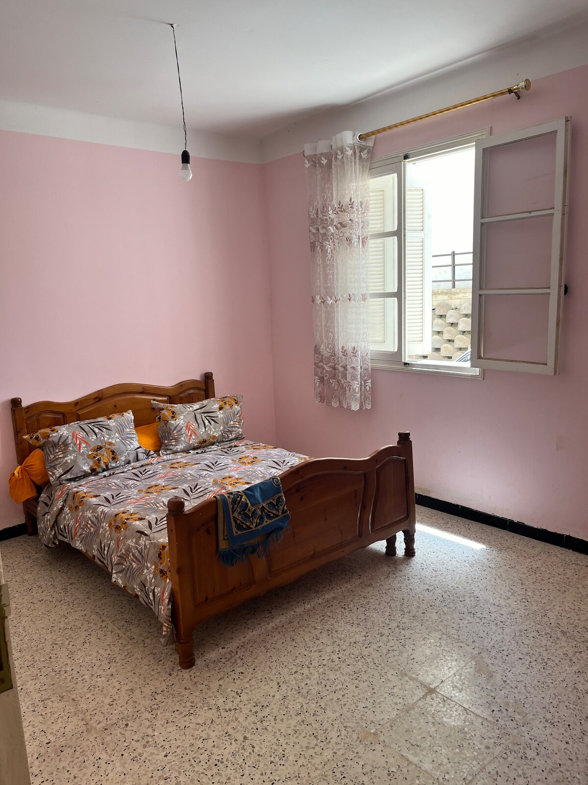 A cozy sleeping area features a wooden bed with a floral-patterned duvet and orange accents. Light enters through a window flanked by sheer curtains, brightening the pale pink walls and highlighting the tiled floor.