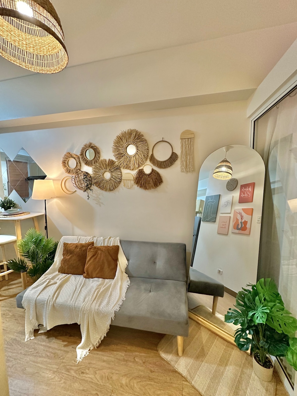 A stylish seating area features a gray sofa adorned with two brown pillows, accompanied by a cozy white throw. Unique wall decor displays various woven mirrors. A large floor mirror reflects the space, while greenery adds a fresh touch to the inviting atmosphere.