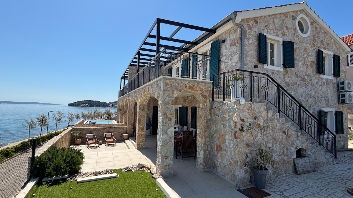 A newly constructed stone house features an outdoor terrace with a sea view. The spacious terrace is equipped with seating and offers easy access to the water. Lush greenery and a pathway lead to the beach, enhancing the peaceful coastal environment.