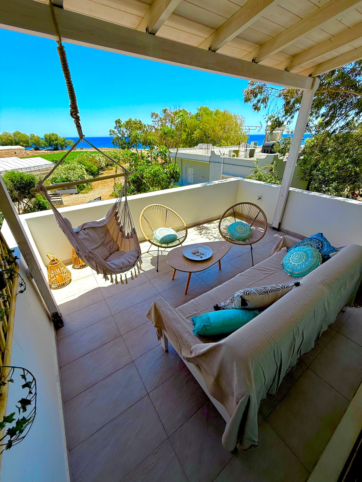 A shaded balcony is furnished with a cozy sofa and two round chairs, offering a relaxing area with colorful cushions. A small table sits at the center, and a hammock is suspended nearby. The view features greenery and the distant sea, enhancing the serene atmosphere.