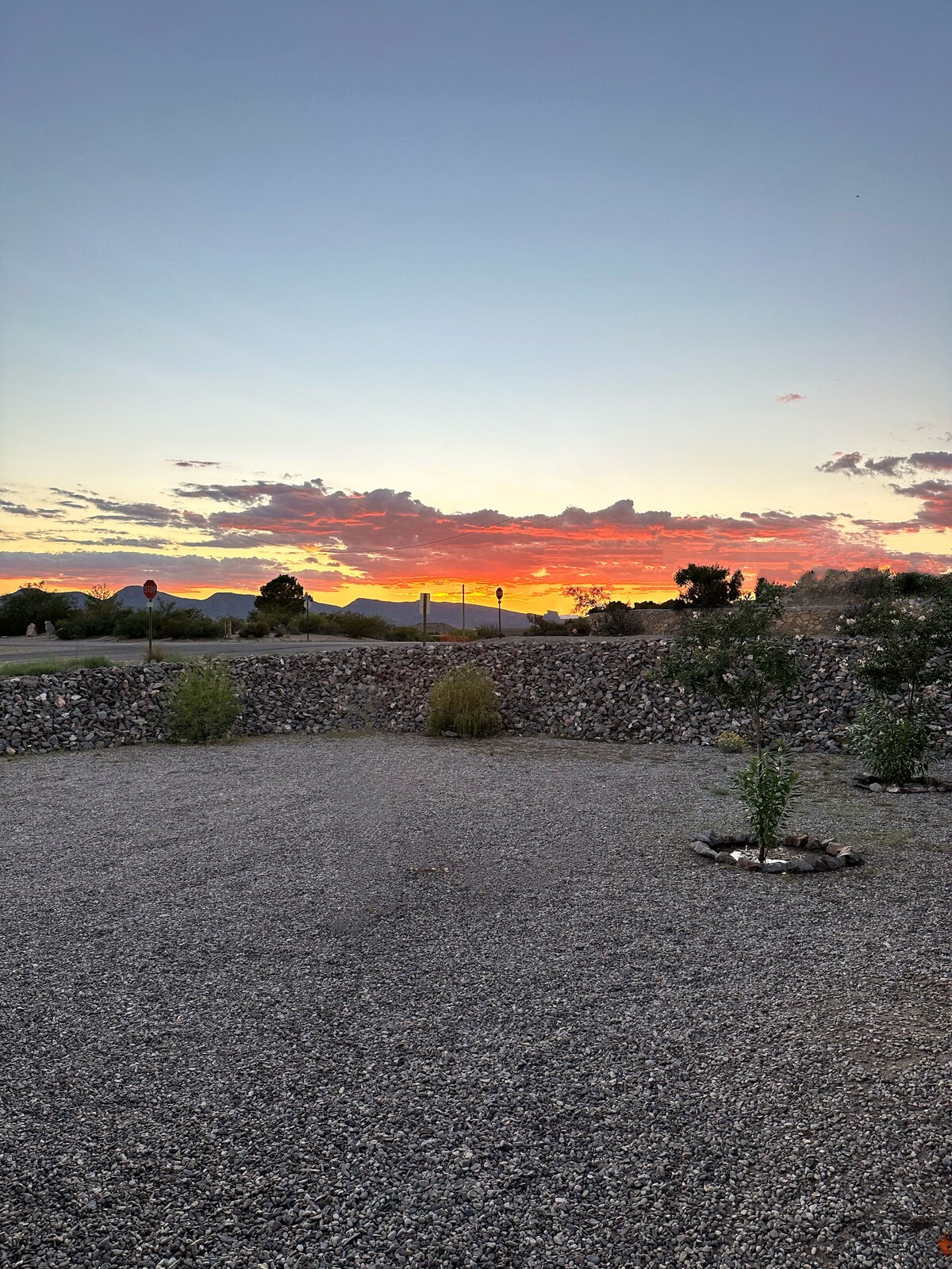 The exterior view captures a stunning sunset with vibrant colors filling the sky. A landscaped gravel area is visible, bordered by low shrubs and rocks, while the distant mountains create a scenic backdrop against the colorful horizon.