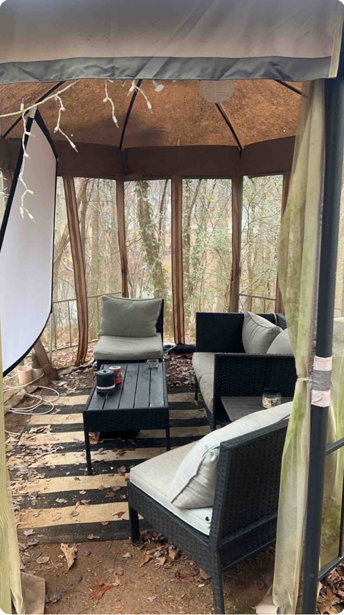 A sheltered seating area is presented with dark wicker furniture, featuring two cushioned chairs and a sleek coffee table. Natural surroundings are visible through sheer curtains, while a projector screen is discreetly positioned for entertainment. Leaves cover the ground, indicating a connection to the outdoor space.