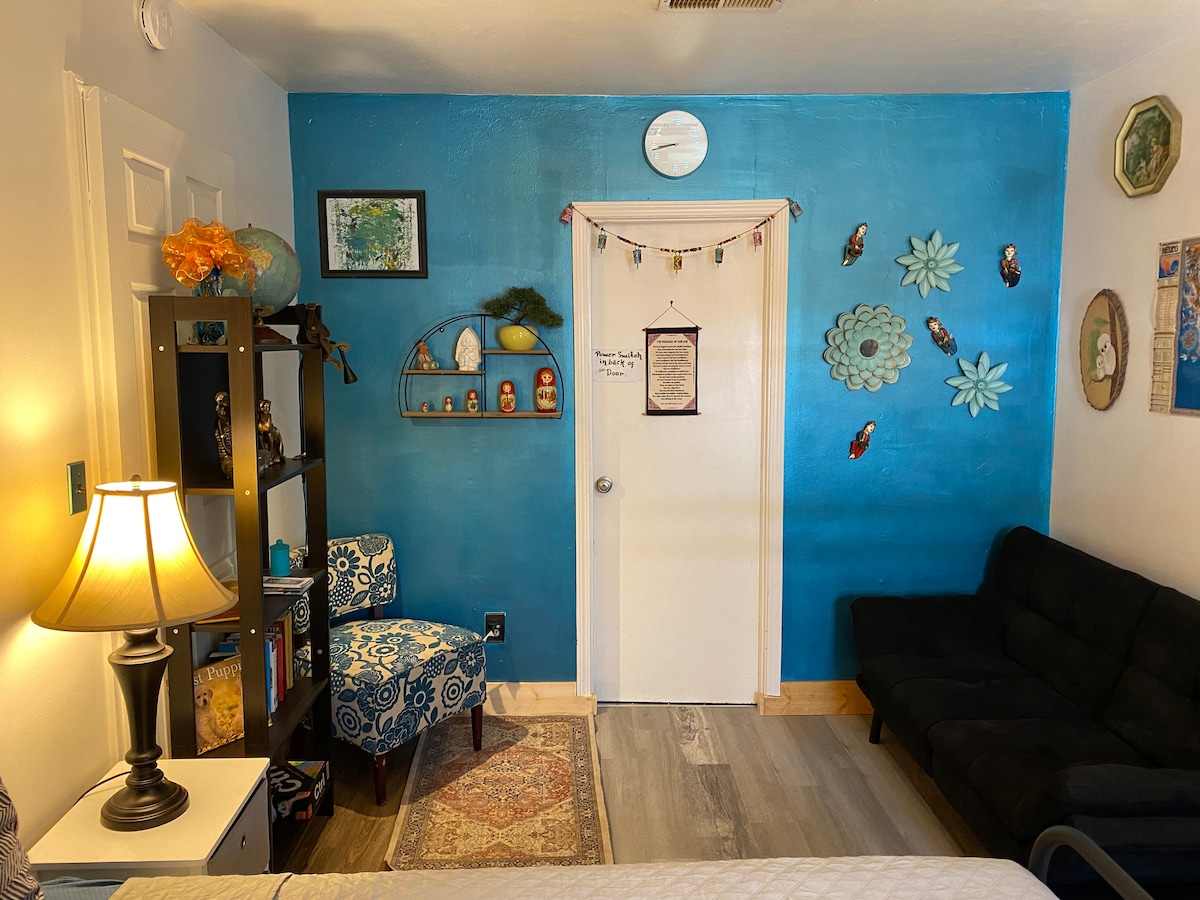 A vibrant room features a striking blue accent wall adorned with decorative wall art and shelves. A cozy chair is positioned near a lamp, while a sofa bed offers additional seating. Natural light illuminates the space, enhancing the cheerful atmosphere.