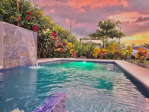 A serene private pool is surrounded by lush tropical plants and vibrant flowers. A waterfall feature adds a soothing element as colors of the sunset reflect in the water, enhancing the peaceful atmosphere of the outdoor space.