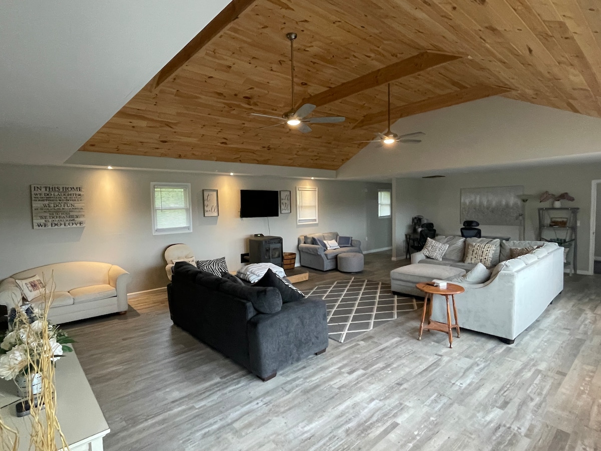 غرفة معيشة كبيرة الحجم تزيد مساحتها عن 700 قدم مربع تتسع لـ 12 شخصًا بشكل مريح وتحتوي على أريكة صغيرة قابلة للسحب مقابل الجدار وسقوف صنوبر جميلة ومروحتان للسقف. جدار مجاور لمحطة العمل