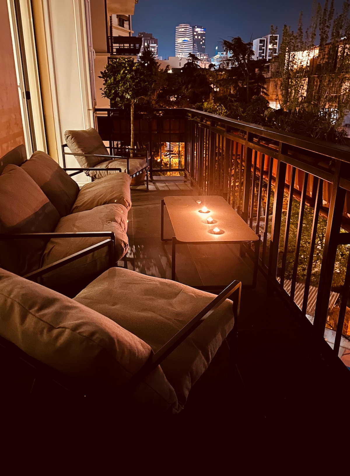 A spacious balcony is illuminated by soft candlelight, featuring two comfortable chairs and a table. Potted plants add a touch of greenery, while the city skyline is visible in the background, creating a serene outdoor space for relaxation.