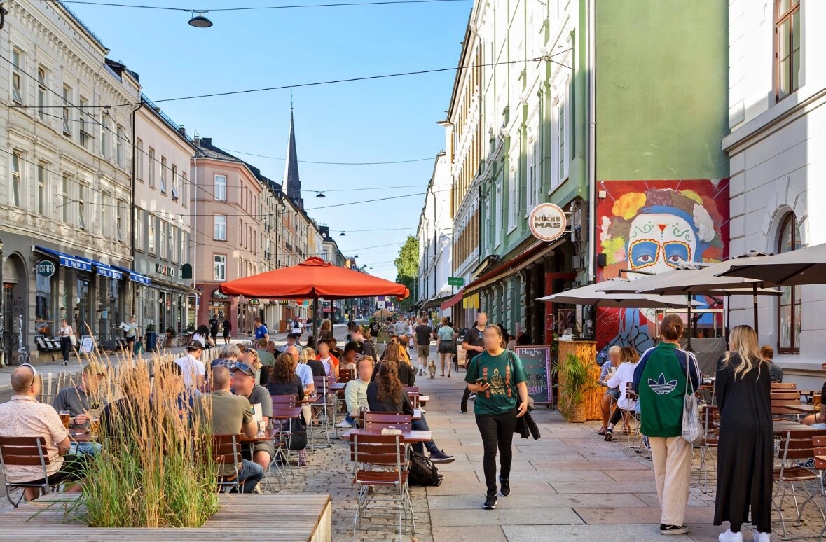 A lively street scene in Grünerløkka features people enjoying the outdoors at tables beneath large umbrellas. Colorful murals adorn nearby buildings, while planters with tall grasses add a natural touch. The street is lined with shops and cafes, creating a vibrant atmosphere.
