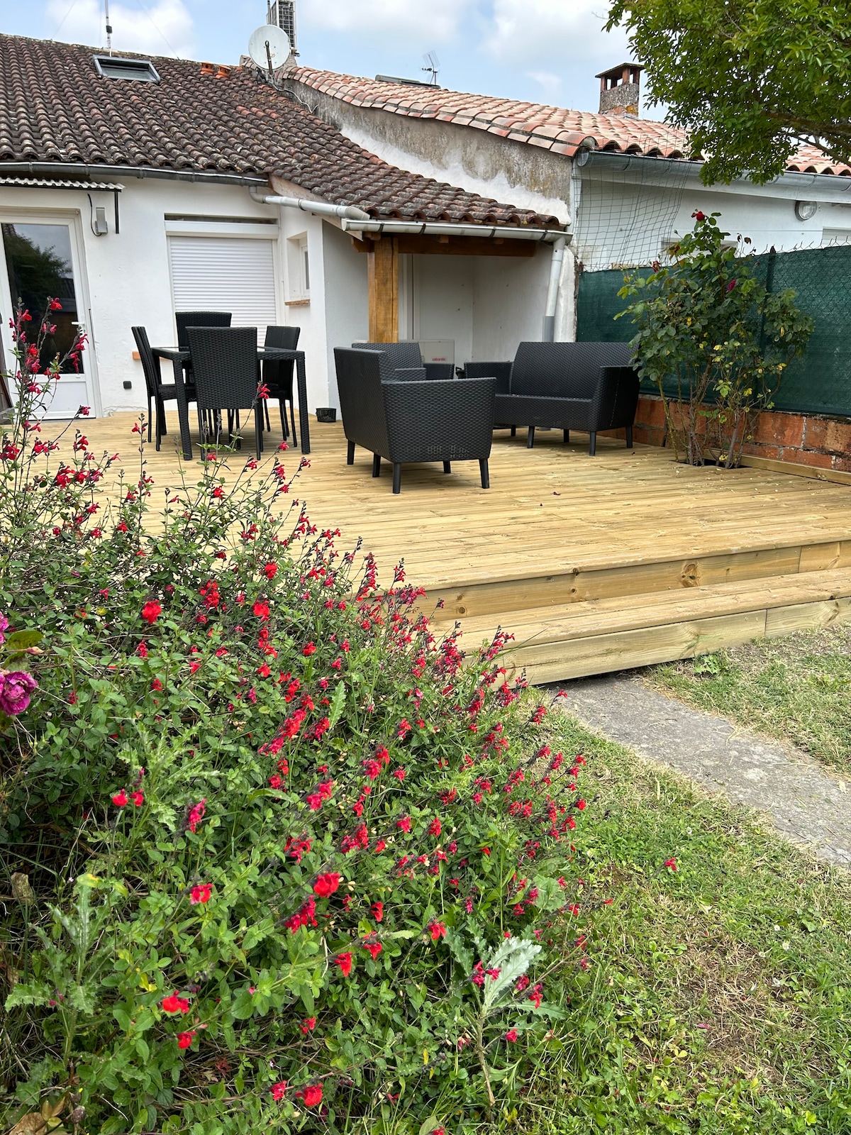 A wooden deck area is featured, furnished with a dining table and several chairs. Colorful flowering plants surround the space, enhancing the greenery. In the background, a shaded area is visible, offering a cozy spot for relaxation.