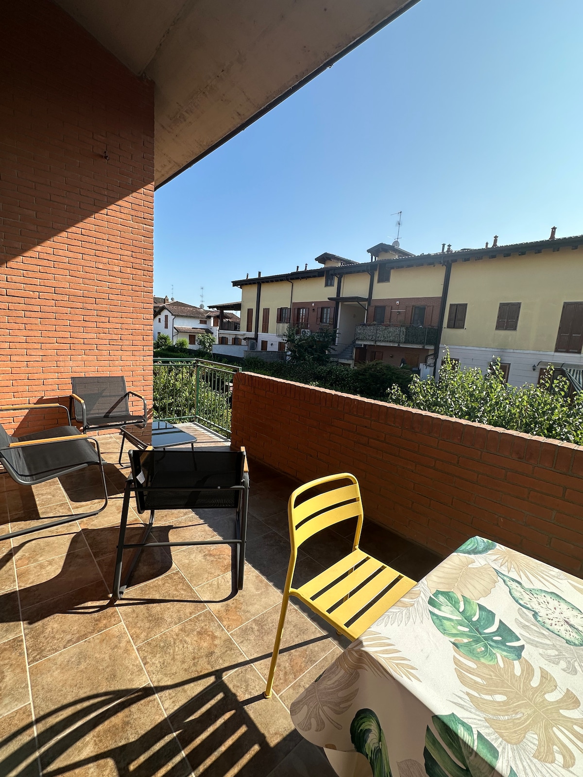 A balcony area is captured with a mix of outdoor seating, including a yellow chair and two black chairs. A table dressed in a patterned tablecloth provides a space for relaxation. Sunlight bathes the space, highlighting the surrounding views of residential buildings and greenery.