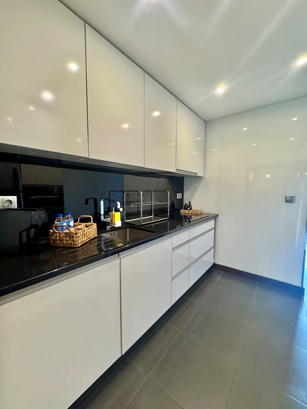 The kitchen space is characterized by sleek cabinetry with a high-gloss finish. A black countertop is paired with modern appliances, including a coffee maker and storage baskets. Bright lighting enhances the clean lines and minimalistic design throughout the area.