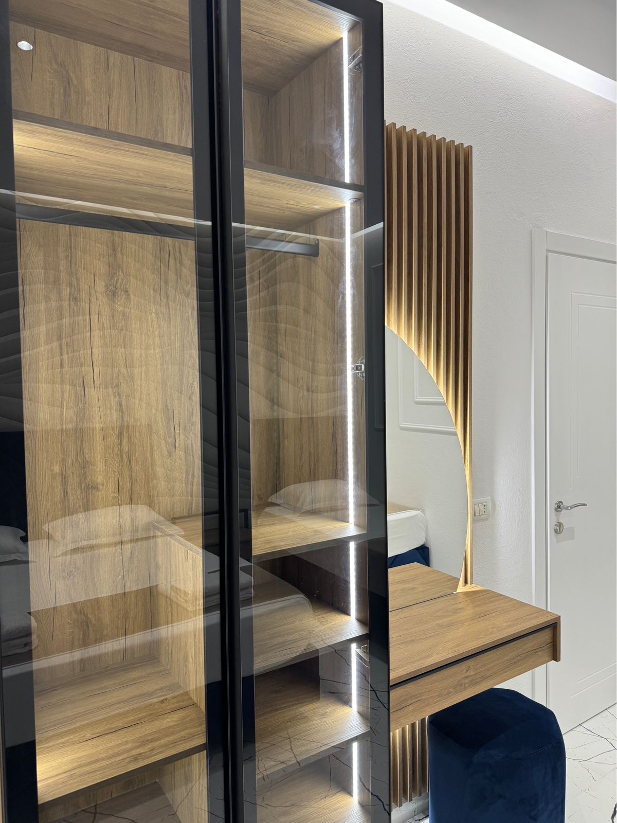 A modern storage unit is visible with a combination of glass and wooden shelves, softly illuminated by integrated lighting. A comfortable desk area is adjacent, featuring a blue upholstered chair, enhancing the functional space for relaxation or work.