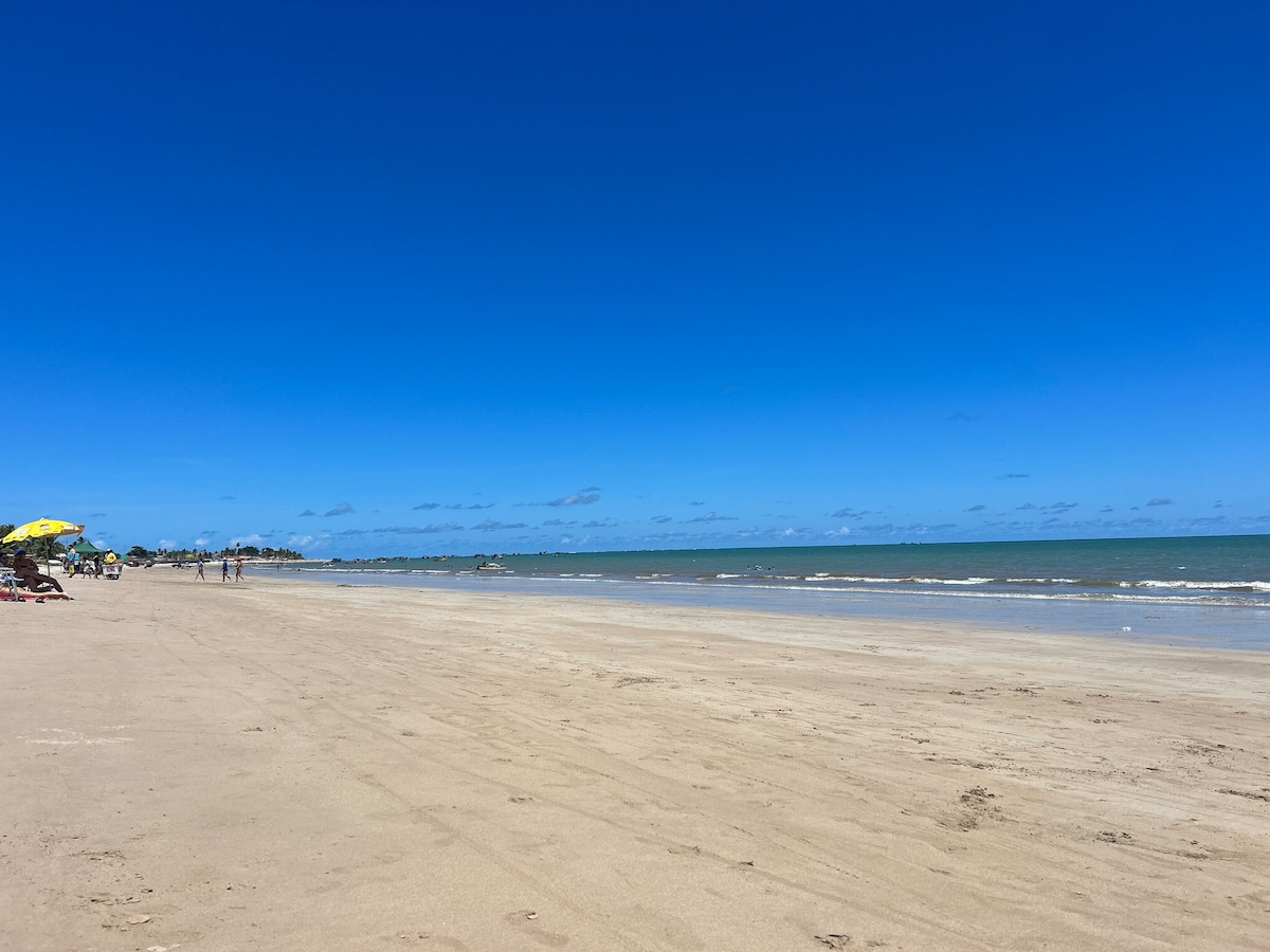A serene beach landscape is depicted, featuring a wide expanse of soft sandy shore under a clear blue sky. Gentle waves lap at the water's edge, while a few umbrellas and beachgoers can be seen in the distance enjoying the vibrant surroundings.
