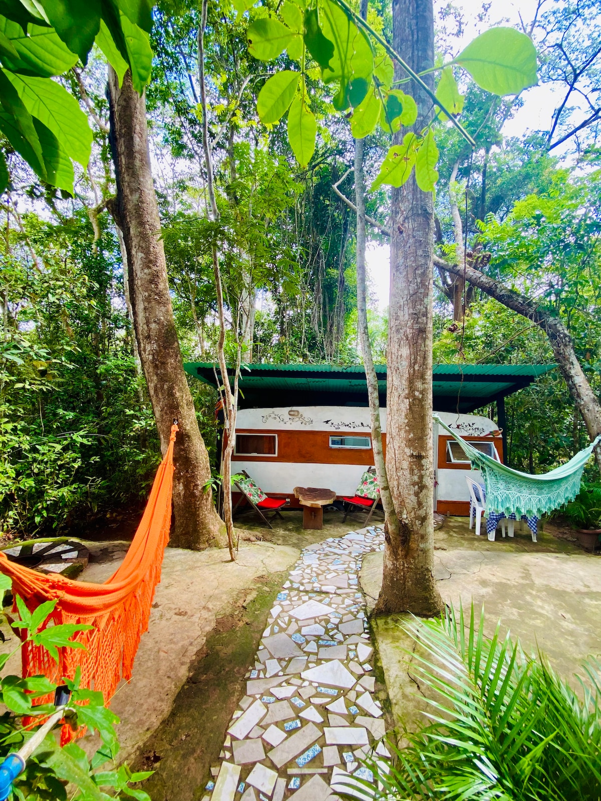 A pathways lined with mosaic tiles leads to a charming trailer nestled among tall trees. Colorful hammocks are suspended nearby, providing inviting spots for relaxation. Lush greenery surrounds the area, offering a serene connection to nature.