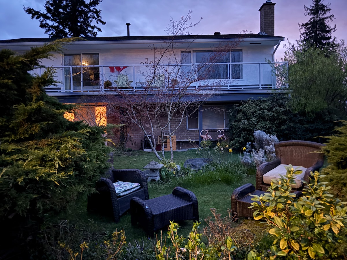 An inviting outdoor seating area is surrounded by lush greenery. Comfortable wicker chairs are positioned among a garden that includes various plants and flowers. The home’s upper level glows softly with warm light, highlighting its balcony that overlooks the serene landscape.
