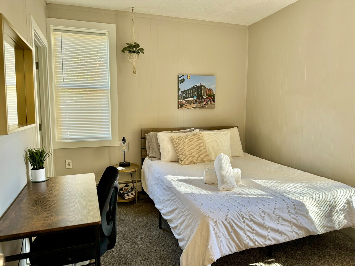 A bright and inviting bedroom is featured, with a spacious bed covered in crisp white linens. A small desk sits nearby, accompanied by a chair. A wall-mounted plant adds a touch of greenery, while a window allows natural light to illuminate the space.
