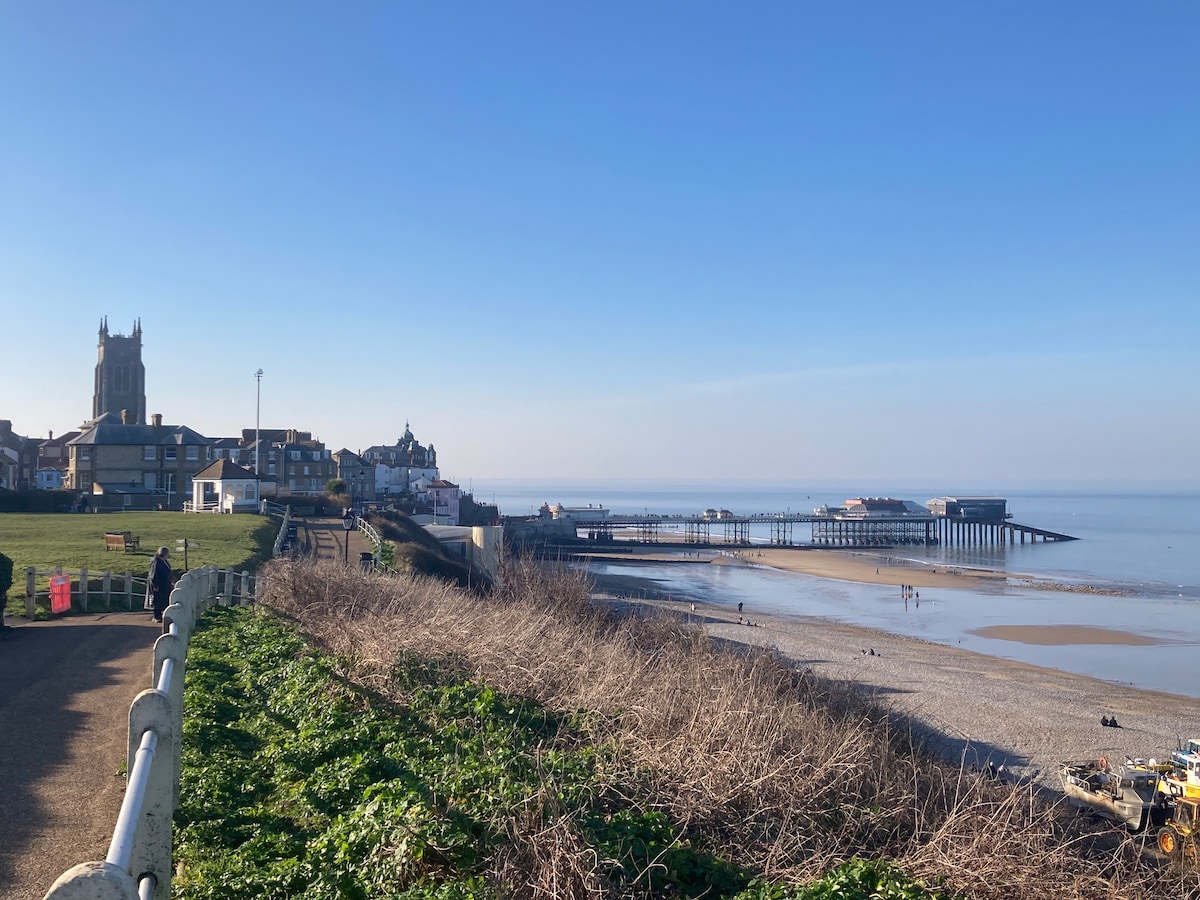 The image captures a panoramic view of the beach alongside a coastal town. The shoreline features gentle waves and a long pier extending into calm waters. Buildings are visible in the background, with a church tower standing prominently. A clear blue sky provides a serene backdrop.