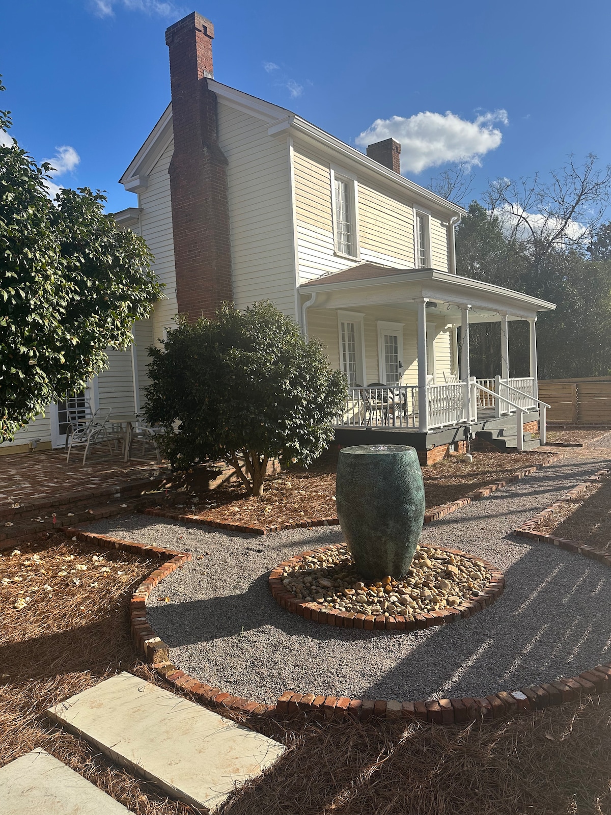 A charming two-story house is framed by greenery, with a front porch featuring white railings. A circular pathway composed of gravel and brick leads to a decorative blue-green urn surrounded by pebbles, enhancing the property's outdoor appeal.