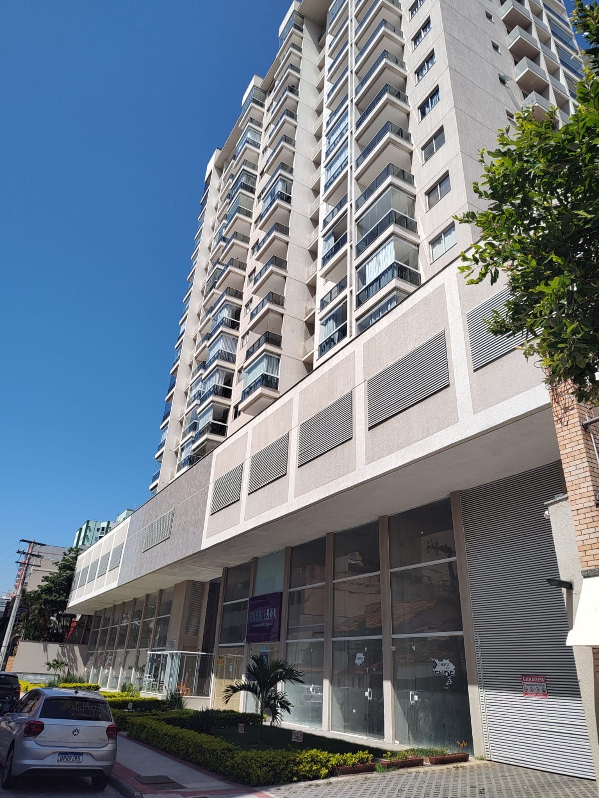 The exterior of the building is depicted, showcasing a modern high-rise structure with multiple floors and large windows. A landscaped area with greenery is visible at the base, and the environment appears sunny and inviting.
